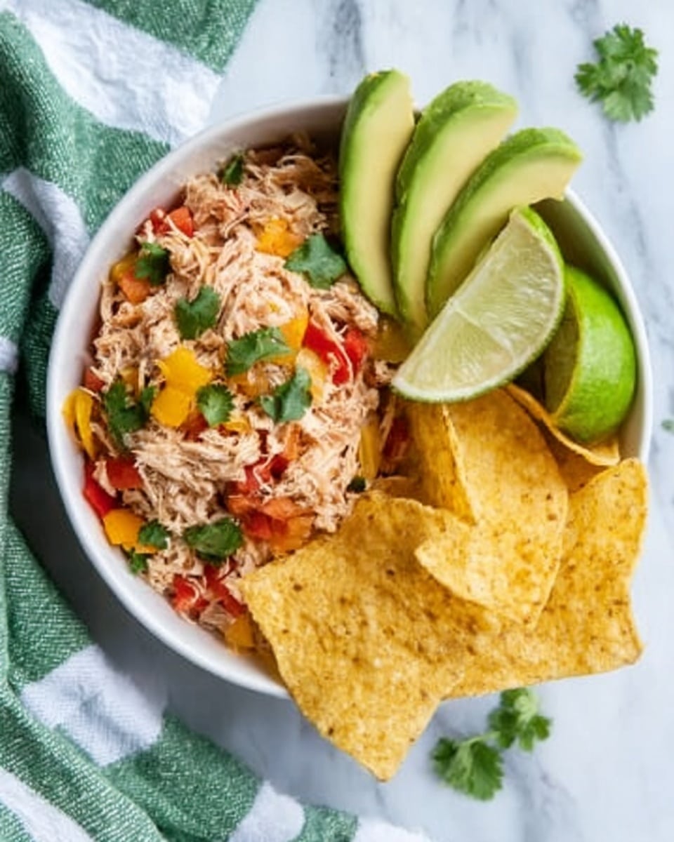 A white bowl on a white marbled surface holds a colorful layered dish. On the left side, there is shredded chicken mixed with red and yellow bell peppers and small bits of onion, all topped with fresh green cilantro leaves. Next to it are thick, curved slices of bright green avocado fanned out. On the right side, a stack of golden tortilla chips creates a crunchy texture. A wedge of lime sits near the shredded chicken, adding a fresh touch. The bowl is placed on a white marbled surface with a green and white striped cloth nearby. photo taken with an iphone --ar 4:5 --v 7