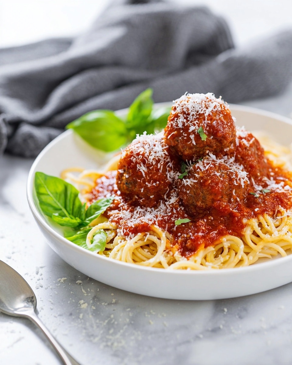 A white deep plate holds a neat pile of spaghetti at the bottom, pale yellow and slightly shiny with a smooth texture. On top of the spaghetti, a rich red tomato sauce covers it well. There are five round meatballs placed in a pyramid shape, covered fully in the same red sauce. The meatballs have a slightly rough texture under the sauce, showing a browned meat surface. Fresh green basil leaves with a smooth, soft look sit on the left side next to the meatballs. Finely grated white cheese is sprinkled evenly over the meatballs and sauce, adding a light, fluffy texture. The plate is set on a white marbled surface, and in the left bottom corner, the tip of a shiny silver fork is visible. The background is softly blurred with a light tone, hinting at a dark cloth but keeping focus on the dish. Photo taken with an iphone --ar 4:5 --v 7