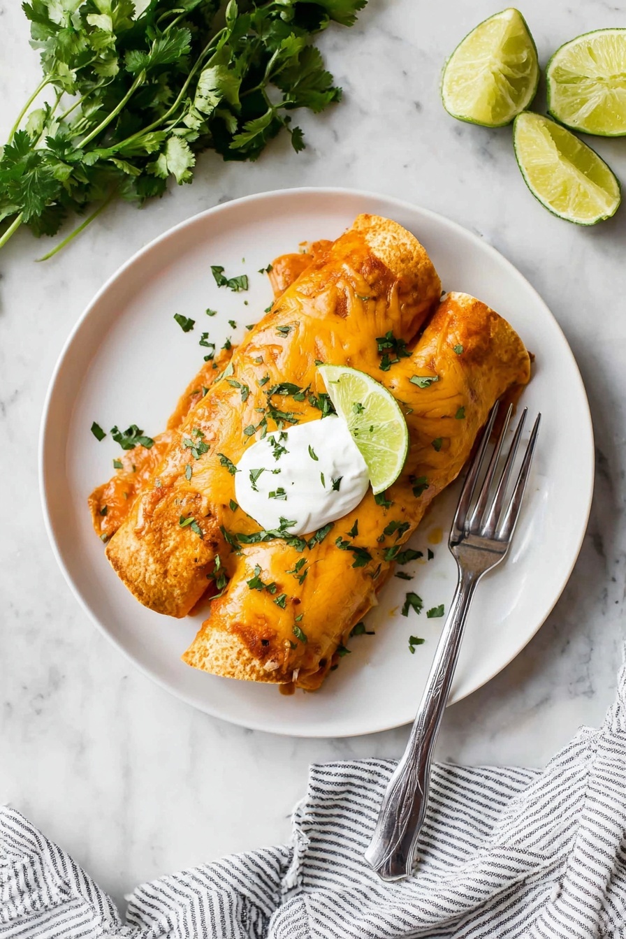 Two rolled tortillas filled and covered in an orange sauce with melted cheese on top are placed side by side in the center of a white plate. A dollop of white sour cream sits on the middle of the tortillas with a slice of lime resting against it. Small green chopped herbs are sprinkled over the dish. A silver fork lies on the right side of the plate. The plate sits on a white marbled surface with fresh green cilantro at the top left and lime wedges at the bottom left. A striped cloth napkin is partially visible at the bottom right corner. photo taken with an iphone --ar 2:3 --v 7 - Easy Chicken Enchiladas, simple chicken enchiladas recipe, quick chicken enchiladas, cheesy chicken enchiladas, flavorful chicken enchiladas