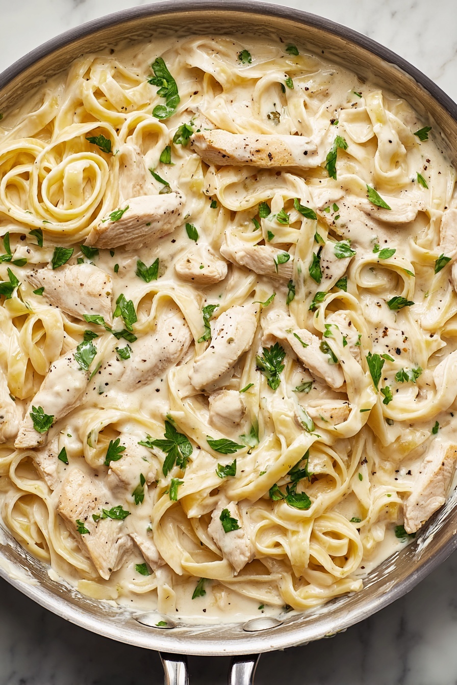 A close-up of a pan filled with creamy white sauce coated fettuccine noodles mixed with sliced white chicken pieces throughout. The thick sauce has a smooth texture with small black pepper flakes. On top, there are scattered bright green parsley leaves adding a pop of color. The pan is round and silver, resting on a white marbled surface. photo taken with an iphone --ar 2:3 --v 7 - Creamy Chicken Alfredo Pasta, easy homemade Alfredo sauce, chicken pasta recipes, quick weeknight dinner ideas, restaurant-style chicken Alfredo