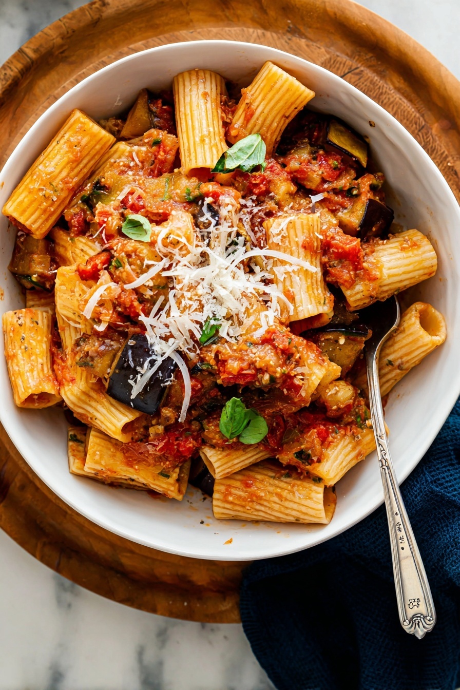 A white bowl filled with rigatoni pasta mixed with chunky red tomato sauce with visible tomato pieces and small dark brown roasted eggplant slices beneath. The pasta and sauce are scattered with fresh green basil leaves and sprinkled with thin, white shreds of cheese. The bowl is placed on a round wooden board on a white marbled surface with a blue and white cloth nearby. A silver fork is inside the bowl on the right side. In the background, part of a red pot with more pasta is slightly visible. Photo taken with an iphone --ar 2:3 --v 7 - Roasted Eggplant Pasta alla Norma, Eggplant pasta recipes, Italian vegetable pasta, easy vegan pasta dishes, hearty veggie pasta