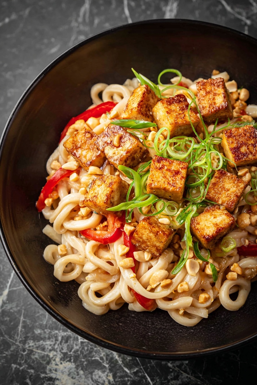 A white bowl filled with a creamy noodle dish showing thick, pale yellow noodles coated in a light brown sauce, mixed with thin green vegetable strips and sprinkled with small bits of peanuts and red chili flakes. There are thin green onion slices layered throughout, adding a fresh contrast. A pair of light wooden chopsticks held by a woman's hand pick up a portion of the noodles, displaying their smooth and slightly glossy texture. The bowl rests on a white marbled surface. In the blurred background, a white bowl containing a dark sauce and chopped peanuts is visible. photo taken with an iphone --ar 2:3 --v 7 - Garlic Peanut Noodles, peanut noodle recipe, spicy Asian noodles, quick vegetarian dinner, creamy peanut sauce noodles