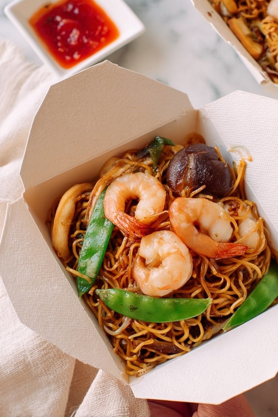 A white round bowl filled with stir-fried noodles layered with orange-colored noodles mixed with pink shrimp, green snap peas, light brown mushroom slices, white bean sprouts, and small red pepper strips, all glistening from the sauce. To the top right, a white takeout box contains a smaller portion of the same noodle dish with three shrimp on top. To the top left, a small white square dish holds a dark red chili sauce with chili flakes. Two bright red chopstick wrappers with gold Chinese writing and a pair of wooden chopsticks rest diagonally on the tray near the sauce dish. All items sit on a white marbled textured surface with a woven white tray underneath most of the dishes. Photo taken with an iphone --ar 2:3 --v 7 - Shrimp Lo Mein, Shrimp Lo Mein recipe, easy shrimp lo mein, homemade lo mein, Asian shrimp stir-fry