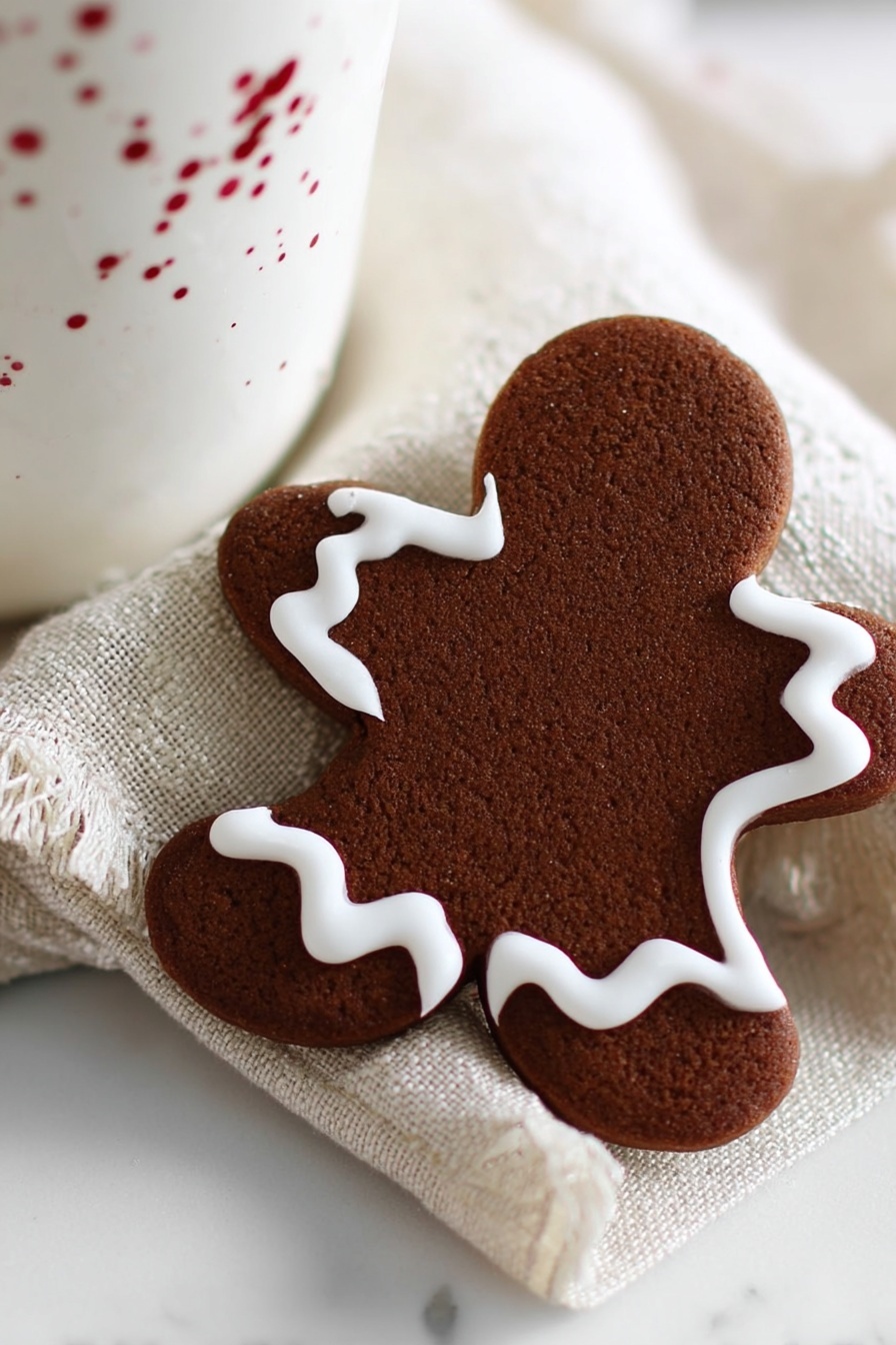 The image shows a single gingerbread cookie shaped like a gingerbread person with one bite taken out of the head. The cookie is a dark brown color and decorated with a simple thick white icing outline that follows the shape of the cookie's edges. It is placed on a beige textured cloth, which lies on a white marbled surface. Part of a white cup with small red speckles can be seen behind the cookie. The overall setting is bright and softly lit. photo taken with an iphone --ar 2:3 --v 7 - Chocolate Gingerbread Cookies, gingerbread cookie recipe with chocolate, easy chocolate-spiced cookies, holiday gingerbread treats, decadent gingerbread cookies