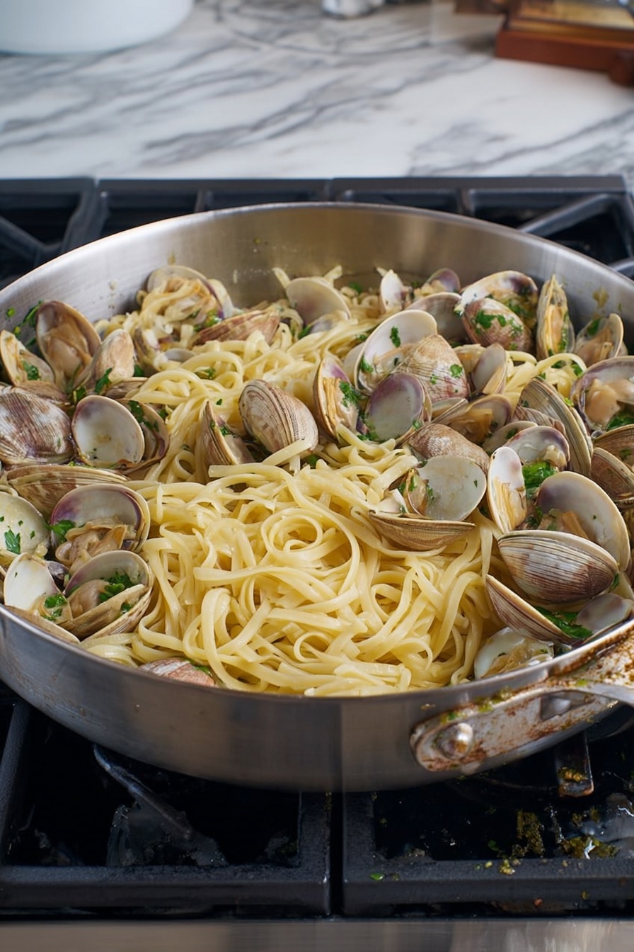 A large silver pan filled with a mix of long, flat, pale yellow noodles and many small clams that have light brown and beige shells, some open showing the inside. The noodles look soft and slightly shiny, tangled with some green parsley pieces scattered on top. The pan rests on a black stove with a white marbled countertop beside it, and some small bits of food cling to the pan's edge. photo taken with an iphone --ar 2:3 --v 7 - Linguine with Clams in White Wine Sauce, clam pasta recipe, seafood pasta with white wine, quick clam linguine, easy clam pasta dish