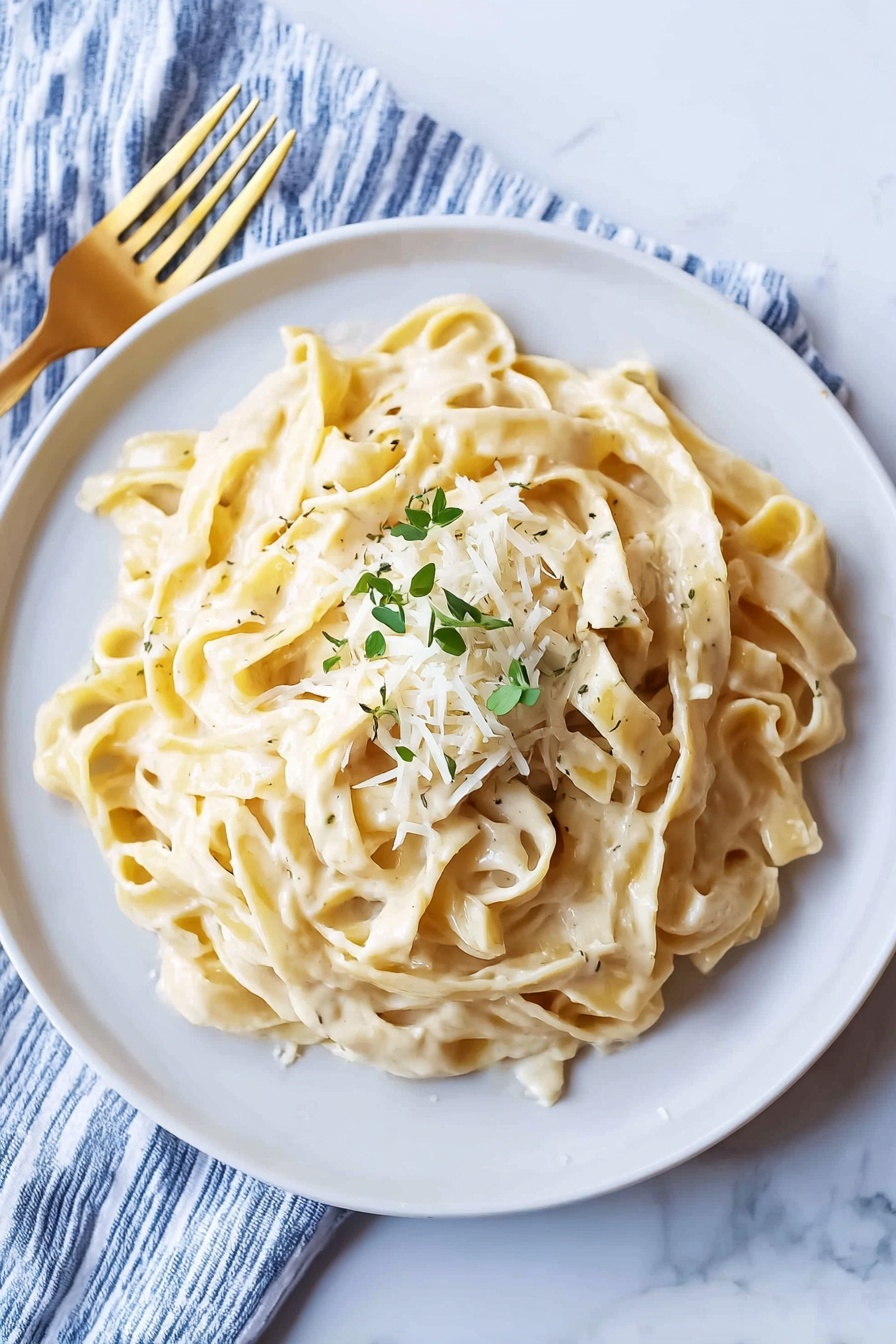 A white round plate holds creamy fettuccine pasta covered in a smooth pale yellow sauce. The pasta ribbons are thick and slightly curled, piled in a loose mound with a small heap of finely grated white cheese and scattered small green herb leaves on top. The plate sits on a white marbled surface next to a blue and white striped cloth napkin, with a golden fork placed to the left. The scene is well lit, highlighting the shiny texture of the sauce. photo taken with an iphone --ar 2:3 --v 7 - Creamy Fettuccine Alfredo, easy Alfredo pasta, cheesy Alfredo sauce, quick pasta dinner, homemade Alfredo recipe