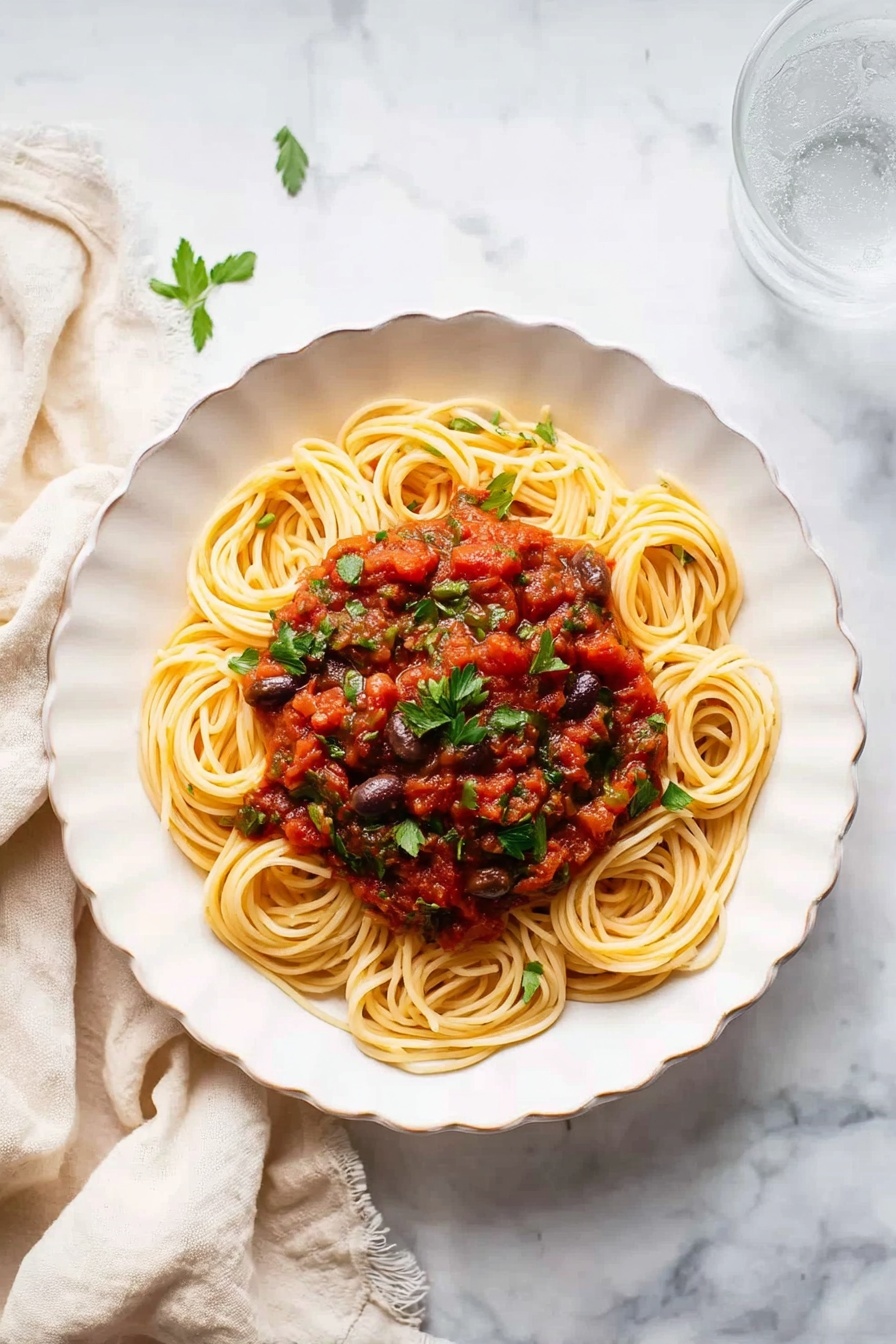 A white plate holds a serving of spaghetti mixed with a chunky red tomato sauce that includes pieces of green herbs and small bits of dark olives or capers. The spaghetti is mostly coated with the sauce, with some pale yellow pasta strands clearly visible, twisted in a loose pile near the center. A silver fork rests on the plate’s edge and is partly wrapped with spaghetti. The plate sits on a white marbled surface next to a clear glass of water and a cream-colored cloth napkin. photo taken with an iphone --ar 2:3 --v 7 - Easy Pasta Puttanesca, Pasta Puttanesca recipe, Italian pasta dinner, quick Italian pasta, briny pasta sauce