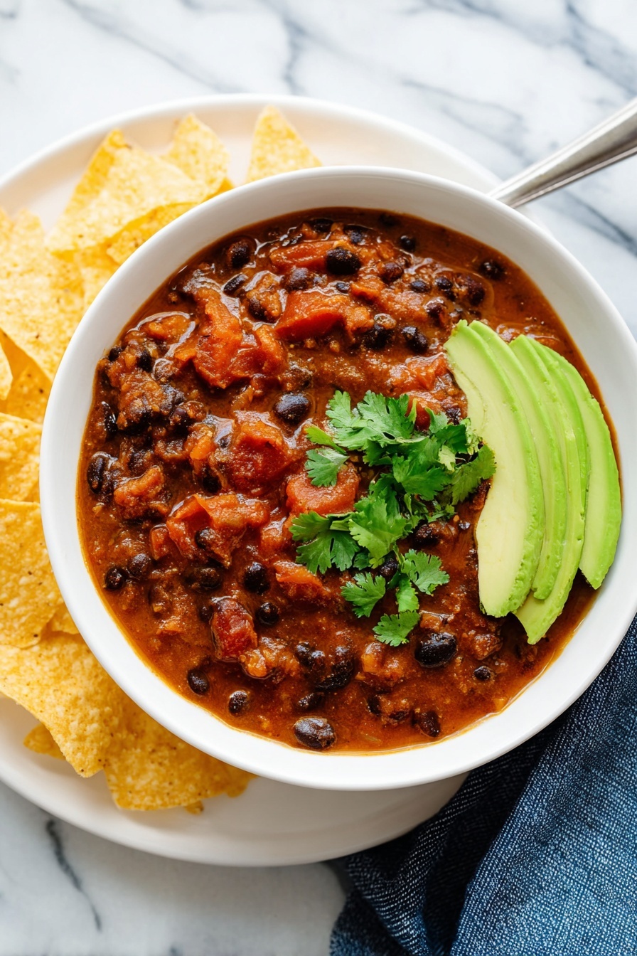 A white bowl filled with thick, chunky chili that has black beans and pieces of tomato in a rich brownish-red sauce, garnished with bright green cilantro leaves. On one side of the bowl, there are thin slices of fresh avocado with a light green color. The bowl sits on a white plate with light yellow tortilla chips arranged around it. The whole setup is on a white marbled surface, and a silver spoon is partially inside the bowl near the avocado. A blue cloth is visible near the bottom right of the bowl. Photo taken with an iphone --ar 2:3 --v 7 - Vegetarian Chili, Vegetarian Chili Recipe, Meat-Free Chili, Healthy Vegetarian Chili, Cozy Vegetarian Chili