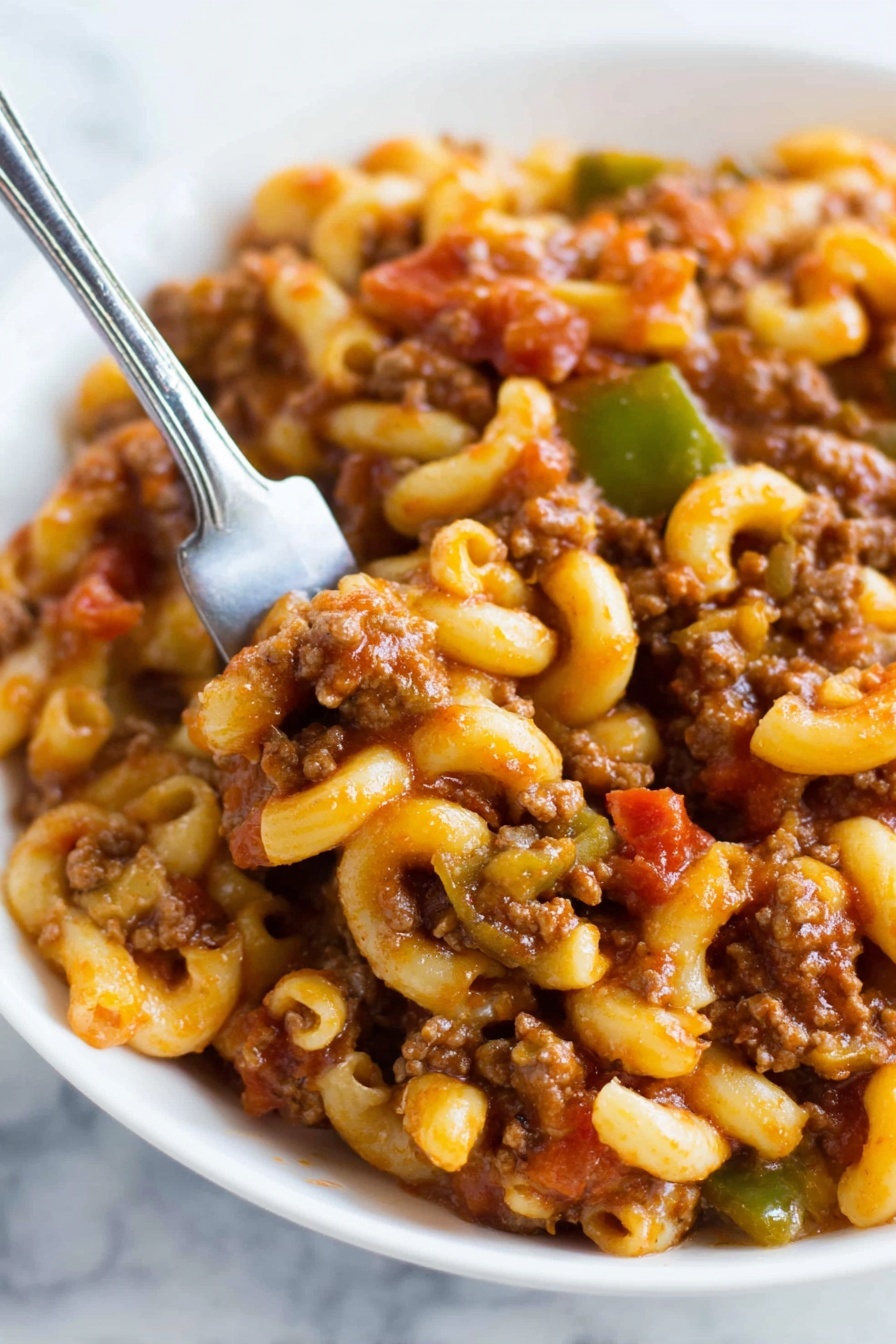 A close-up of a white bowl filled with macaroni and ground beef mix. The dish has elbow-shaped pasta coated with thick, reddish-brown sauce mixed with crumbled browned meat. Small pieces of green vegetables and bits of red tomato are visible through the mixture. A fork with a silver handle is partially inside the bowl, poking into the food. The background shows a white marbled surface. photo taken with an iphone --ar 2:3 --v 7 - American Chop Suey, Easy American Chop Suey, one-pot American Chop Suey, hearty American Chop Suey dinner, simple American Chop Suey recipe