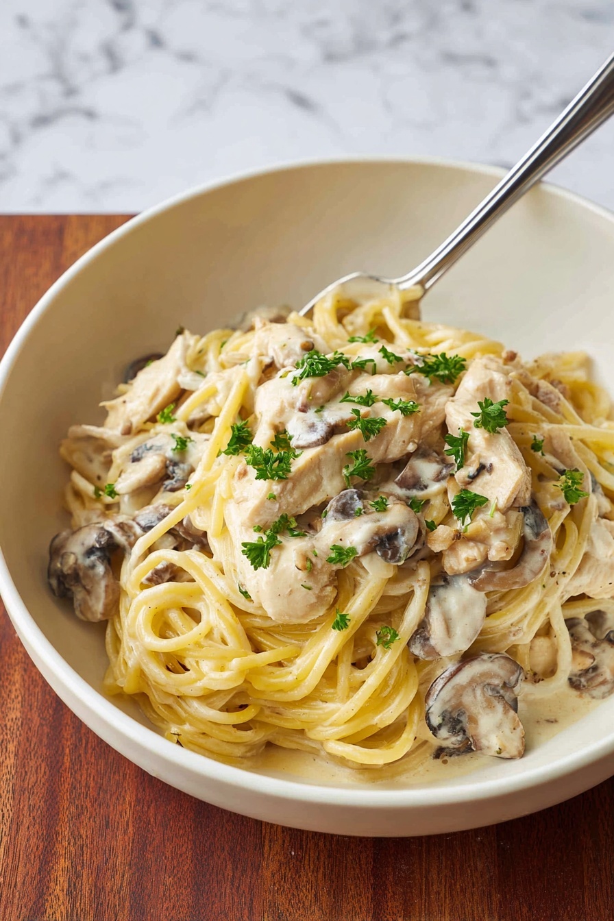 A white bowl holds a creamy pasta dish with about three layers: the bottom layer has pieces of light beige chicken mixed with brown mushrooms in a thick white sauce; the middle layer is covered by long, thick yellow spaghetti noodles twisted over the chicken; the top layer includes small green parsley leaves scattered over the pasta. A silver fork rests on the right side inside the bowl. The bowl sits on a brown wooden surface with a white marbled texture in the background. photo taken with an iphone --ar 2:3 --v 7 - Creamy Chicken Tetrazzini Pasta Bake, easy chicken tetrazzini, cheesy pasta casserole, comforting chicken pasta, homemade tetrazzini recipe