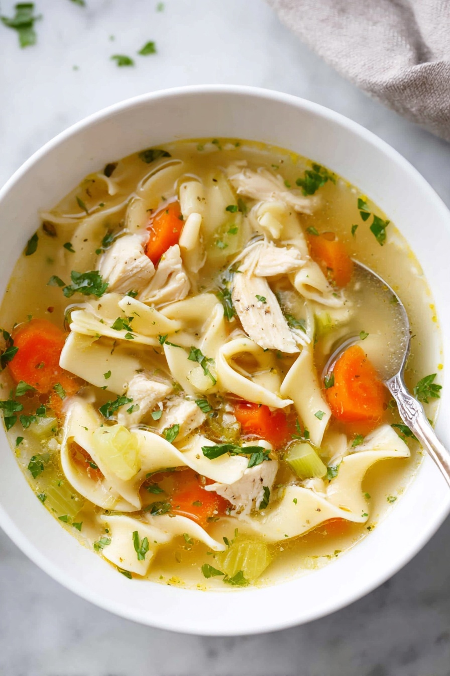 A white bowl is filled with chicken noodle soup showing three main layers: the clear golden broth with tiny green herbs floating, soft wide flat noodles in light yellow twisting through the soup, and bright vegetable pieces including orange carrot rounds and green celery slices scattered along with small pale chunks of white cooked chicken. The bowl sits on a white marbled surface, and there is a shiny spoon in the background. The photo looks fresh and warm, inviting to eat. photo taken with an iphone --ar 2:3 --v 7 - Homemade Chicken Noodle Soup, homemade chicken noodle soup, comforting chicken noodle soup, easy chicken noodle soup, best chicken noodle soup