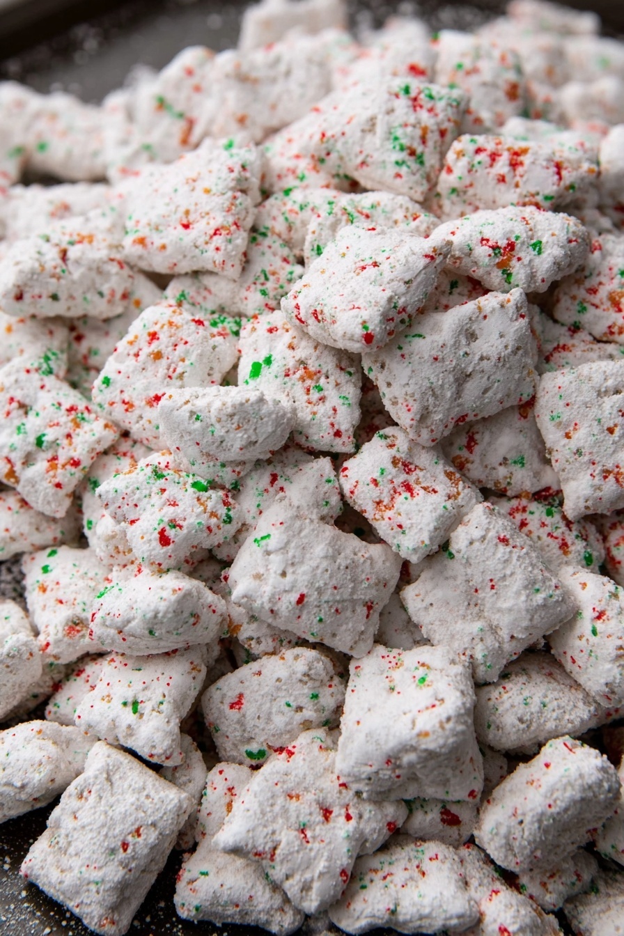 This image shows a large pile of small, irregularly shaped white pieces covered in a powdery coating. The white coating has specks of red and green sprinkled over all the pieces, giving a festive look. The pieces have a rough and uneven texture, suggesting they might be crunchy or dry. They are spread out on a dark tray, but the main focus is the texture and color of the coated pieces. The red and green sprinkles are small and scattered evenly across the surface of each piece, adding a colorful contrast to the white powder coating. photo taken with an iphone --ar 2:3 --v 7 - Festive Chocolate Peanut Butter Puppy Chow, holiday snack mix, easy holiday treats, chocolate peanut butter snack, festive party snacks