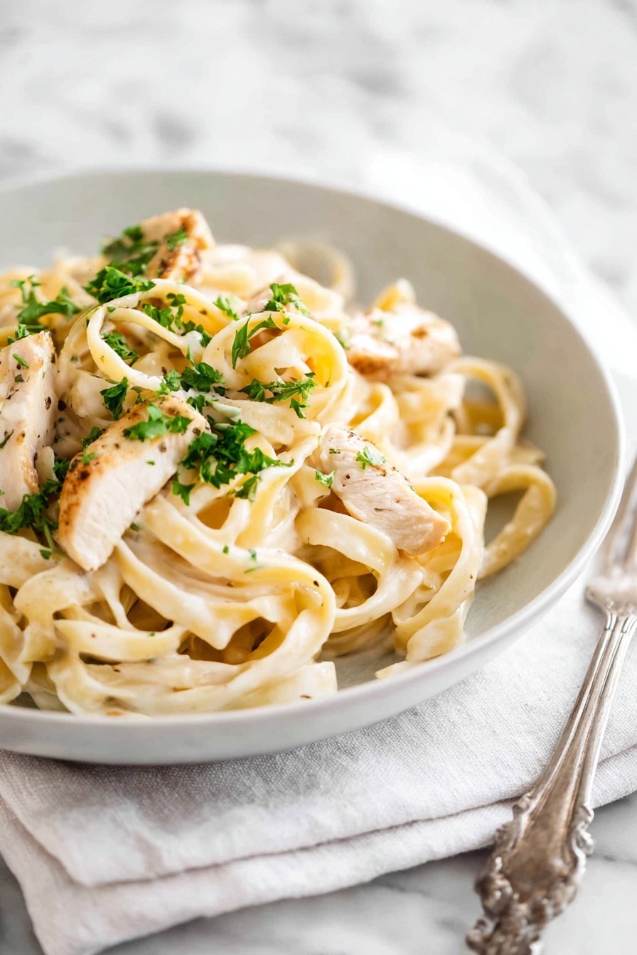A white bowl filled with creamy fettuccine pasta topped with pieces of light golden-brown cooked chicken and sprinkled with small bright green parsley bits. The pasta is coated with a smooth, pale cream sauce and is twisted into soft loops. The bowl sits on a white cloth napkin, all placed on a white marbled surface. A silver fork is positioned to the left side of the bowl. Photo taken with an iphone --ar 2:3 --v 7 - One Pot Chicken Alfredo Pasta, easy chicken Alfredo pasta, quick creamy pasta, one skillet chicken pasta, family-friendly Alfredo dish