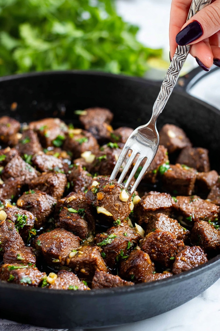 The image shows a black cast iron pan filled with many small chunks of cooked brown meat, each piece sprinkled with bits of green herbs and small pieces of golden-brown garlic. A woman's hand with dark nail polish is holding a silver fork that picks up a single piece of meat from the pan. The background is blurry with green leaves visible, creating a fresh feel. The pan sits on a white marbled surface. photo taken with an iphone --ar 2:3 --v 7 - Garlic Butter Steak Bites, steak bites recipe, easy steak bites, quick dinner ideas, juicy steak tips