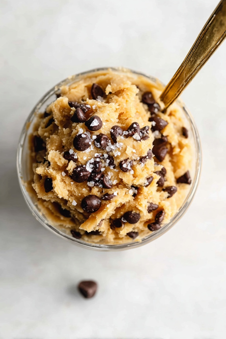 The image shows a clear glass cup filled with a rough-textured mixture that looks like cookie dough. The dough is light beige with many dark brown chocolate chips scattered throughout every layer. The dough is piled high above the rim of the cup in a mound-like shape, with a sprinkling of coarse salt on top. A long silver spoon is stuck into the mound, leaning slightly to the right. The background is a white marbled texture with soft lighting, making the dessert stand out clearly. Photo taken with an iphone --ar 2:3 --v 7 - Vegan Cookie Dough Bites, vegan cookie dough snack, healthy vegan treats, no bake vegan dessert, vegan sweet bites