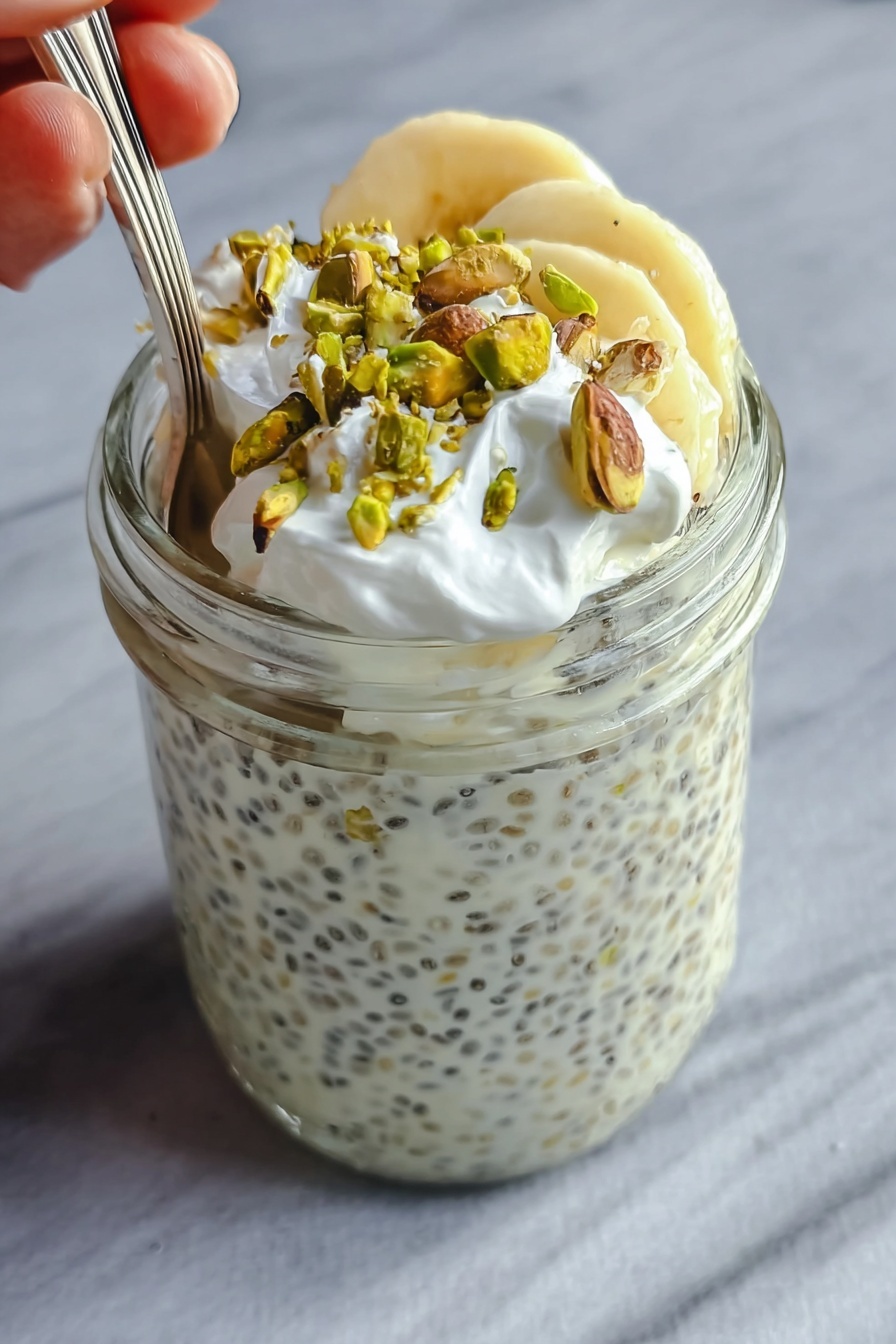 The image shows a clear glass jar filled almost to the top with a creamy white chia pudding that has small black chia seeds spread evenly throughout. On the top layer inside the jar, there are a few slices of light yellow banana, which are partially covered by the creamy pudding. Over the bananas, there is a layer of small white whipped cream dollops, adding a soft texture. Scattered on the very top are roughly chopped green pistachio nuts that add a contrasting color and crunchy texture. A spoon is placed inside the jar, with a woman's hand holding it gently from the left side. The jar is set against a smooth, white marbled surface. Photo taken with an iphone --ar 2:3 --v 7 - Creamy Pistachio Overnight Oats with Protein, pistachio overnight oats, healthy breakfast oats, protein-packed overnight oats, gourmet pistachio oats